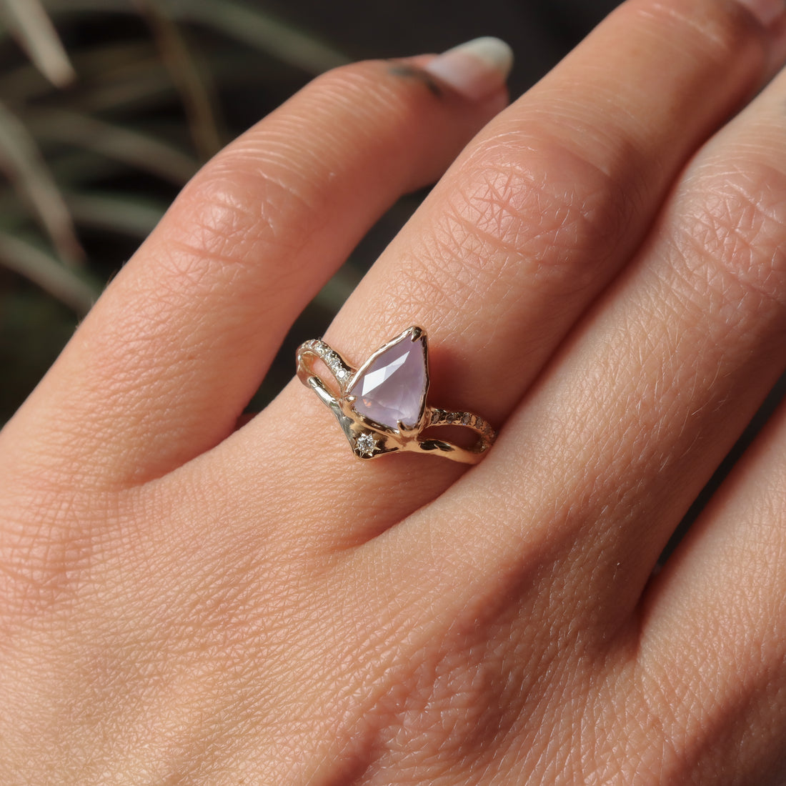 Close-up of a hand wearing a ring with a pink gemstone against a blurred natural background