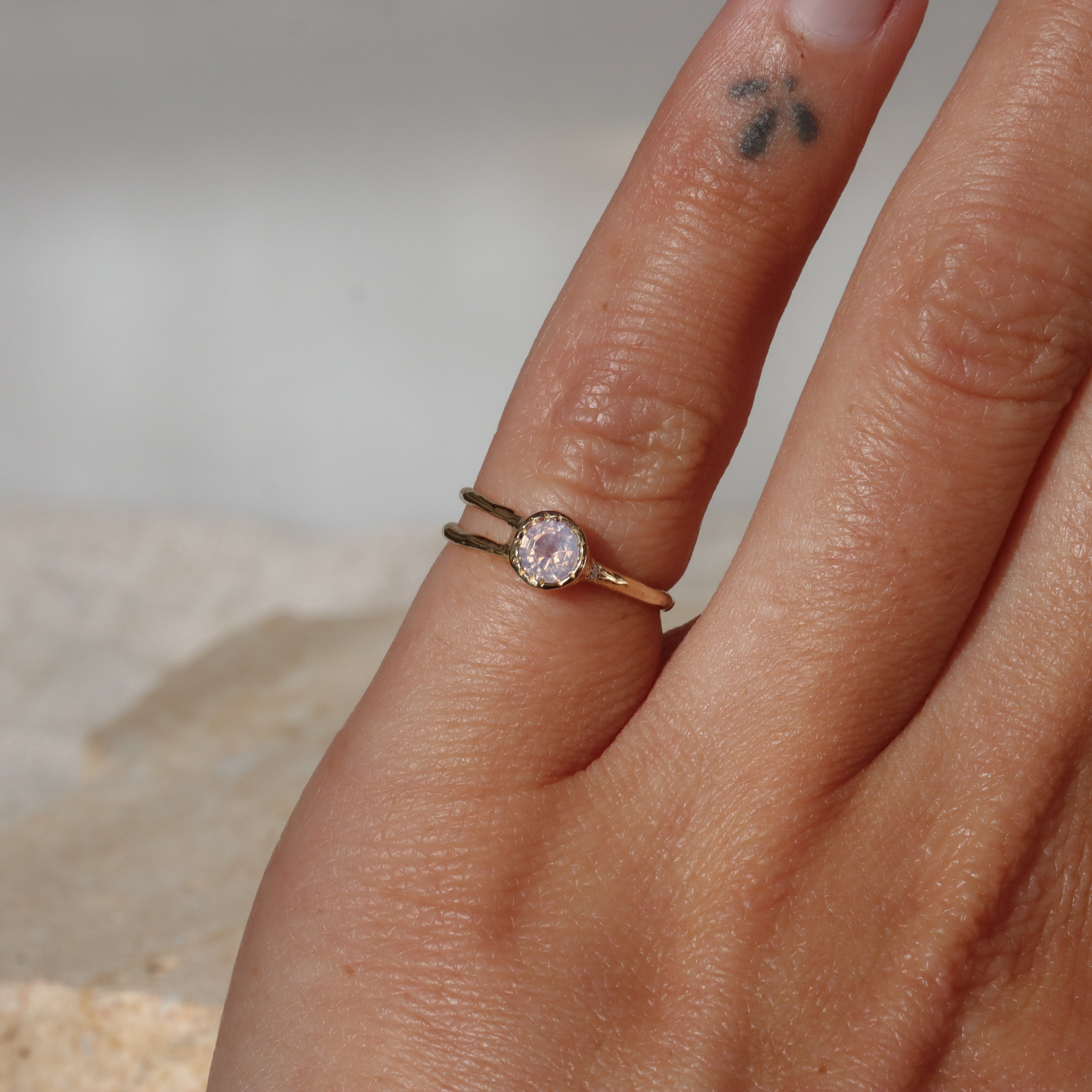 Close-up of a hand wearing a gold ring with a pink gemstone on a neutral background