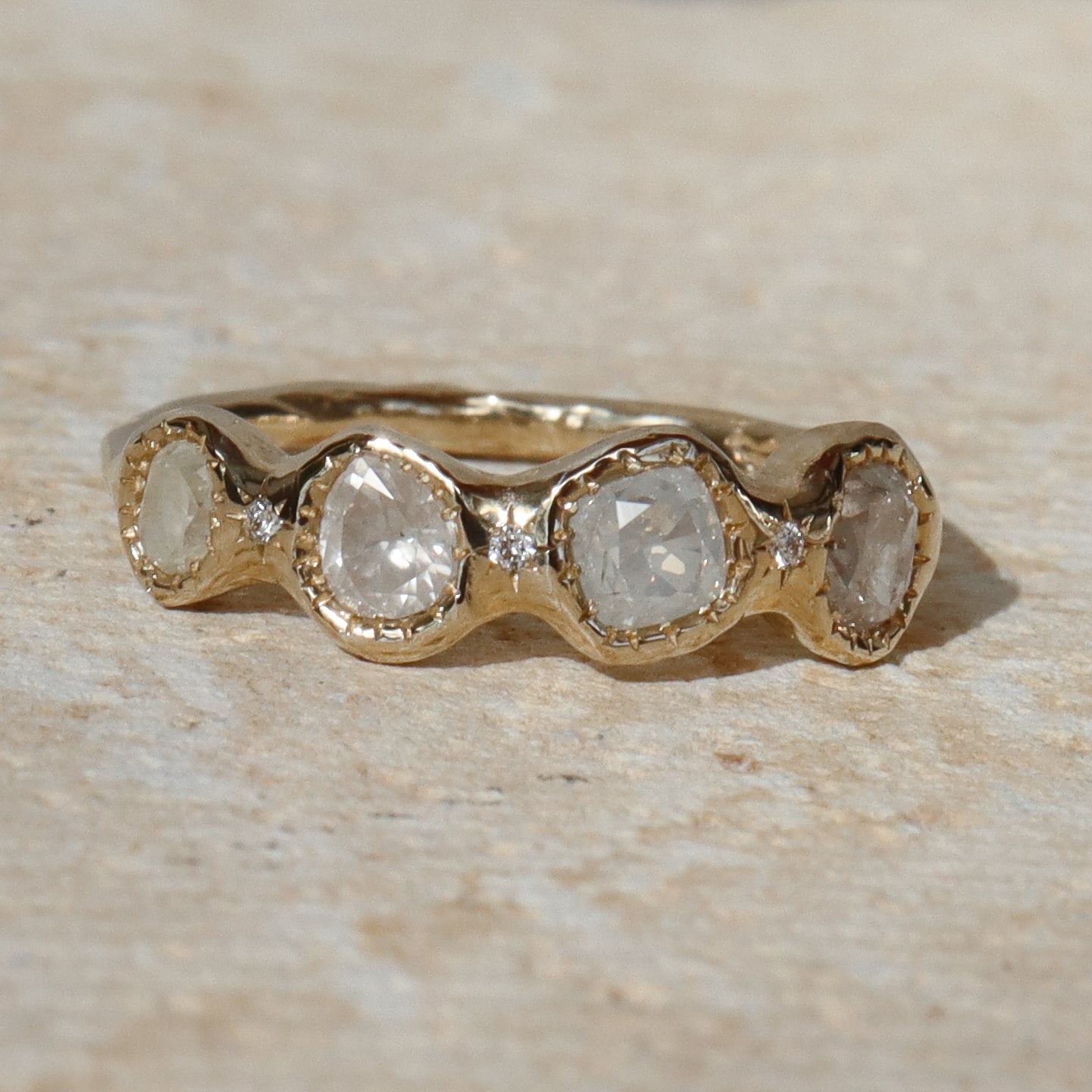 A gold band ring featuring five round-cut clear gemstones in a wavy row, photographed on a light beige stone surface.
