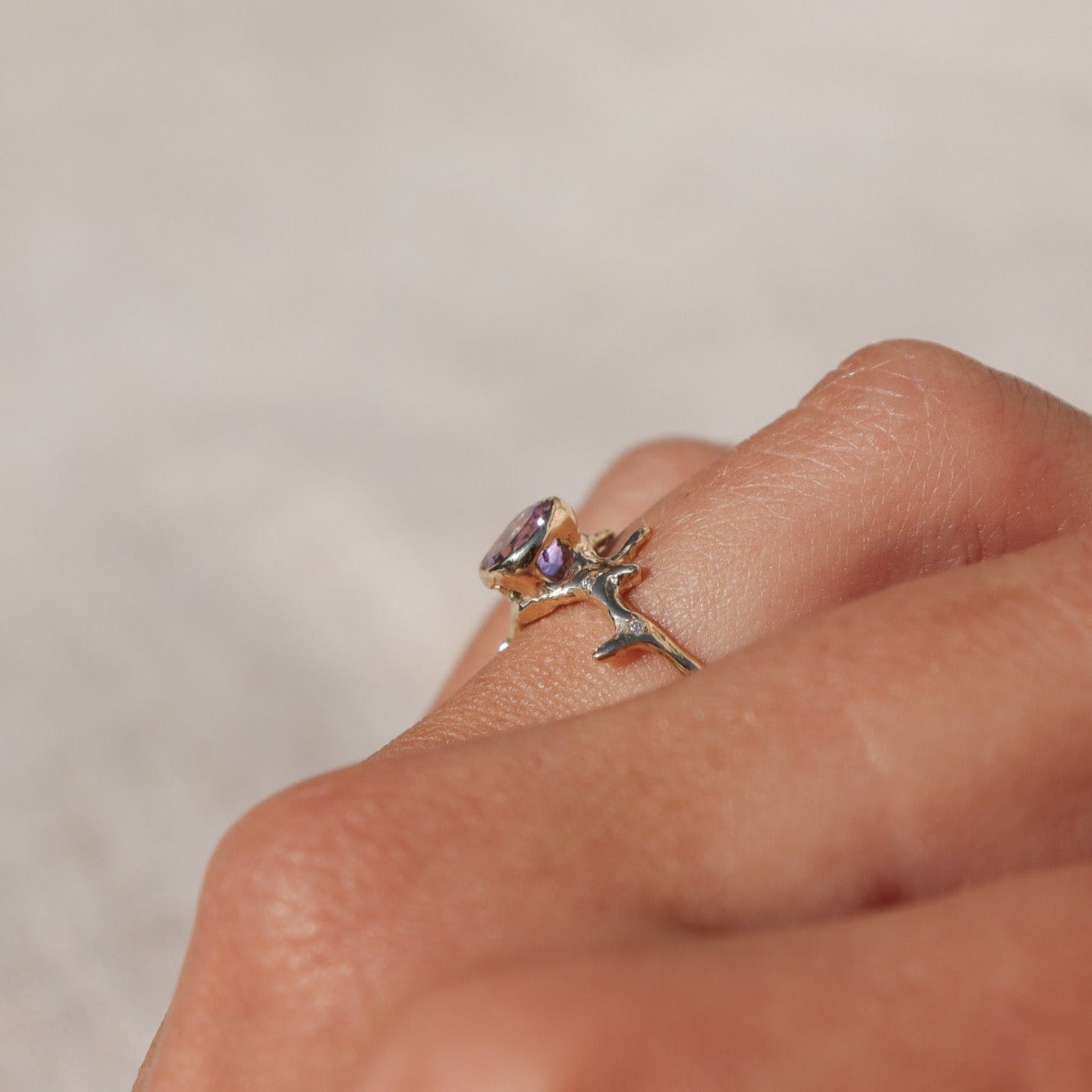Exquisite round brilliant cut ametrine gemstone, bezel-set in a gold band with an organic coral-like design, adorned with sparkling diamond accents, worn on a finger, shown from the side to depict the cut outs under the gem.