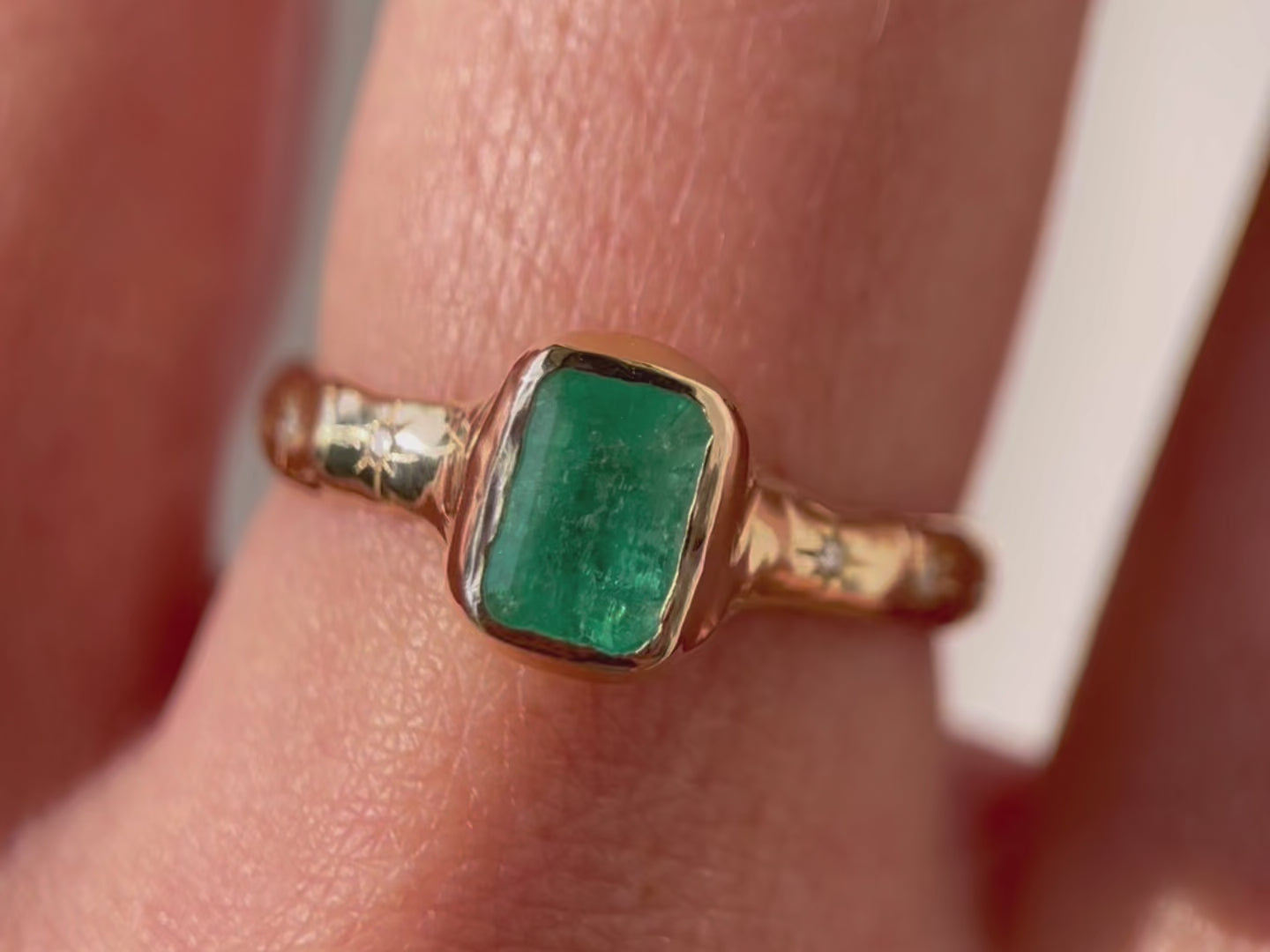 A close up video of an emerald cut emerald ring is bezel set in 14k gold with a wide band that has multiple star set diamonds along the sides.