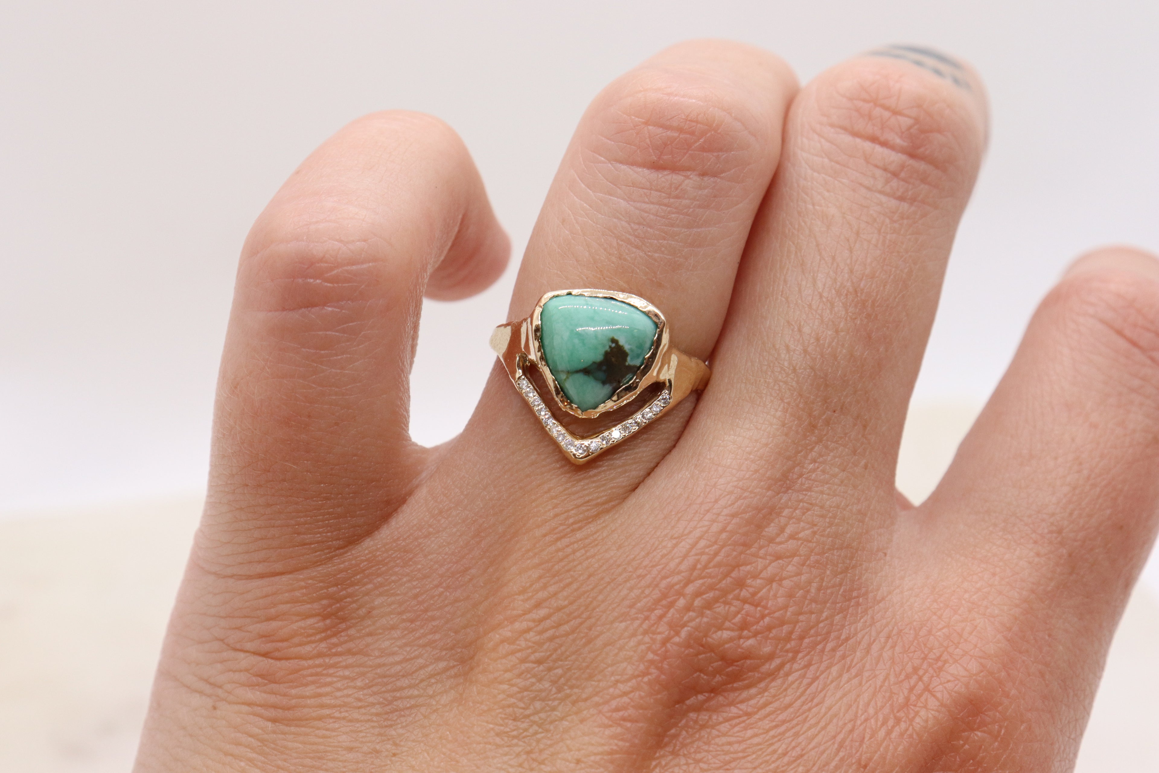 A models hand wears a trillion variscite is bezel set in 14k yellow gold with a v-shape band covered in pave diamonds