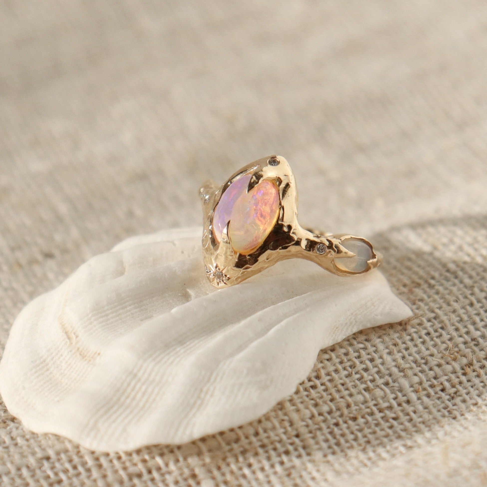 A pink opal is set with prongs in the center of an organically crafted band, there is an oval opal on the side of the band with diamonds on each side.