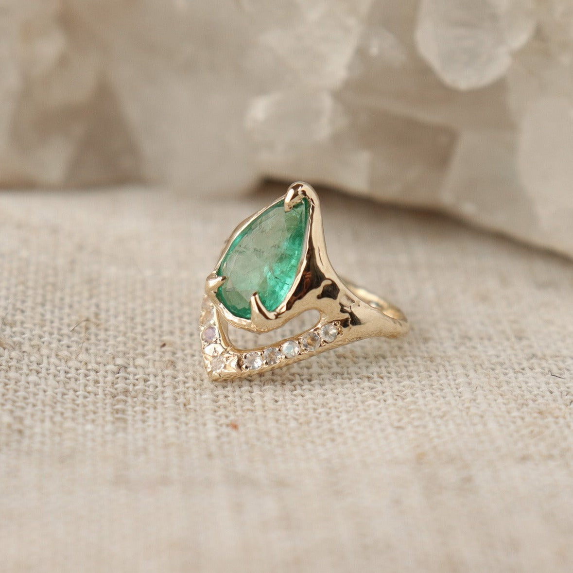 Close up side view of a pear shaped emerald is set with prongs and has a V-shape band under the stone with moonstones.