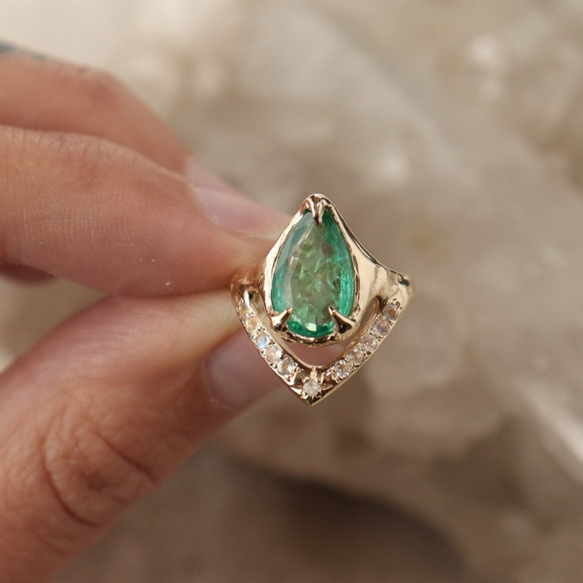 A pear shaped emerald is set with prongs and has a V-shape band under the stone with moonstones.
