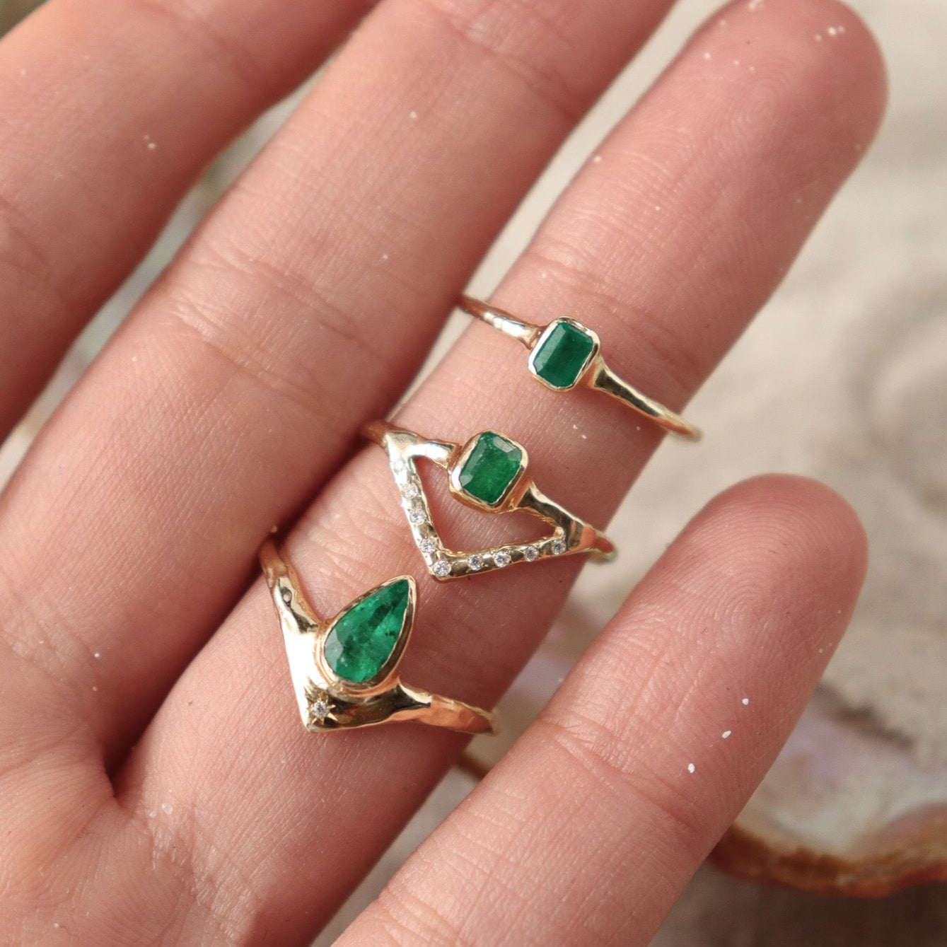 Stunning emerald ring with a bezel-set, rich green gemstone elegantly perched on a delicate, narrow band. Beneath it, a gracefully designed V-band features organically set diamonds, adding a touch of nature-inspired luxury to your jewelry collection. Shown next to two other emerald rings for comparison.