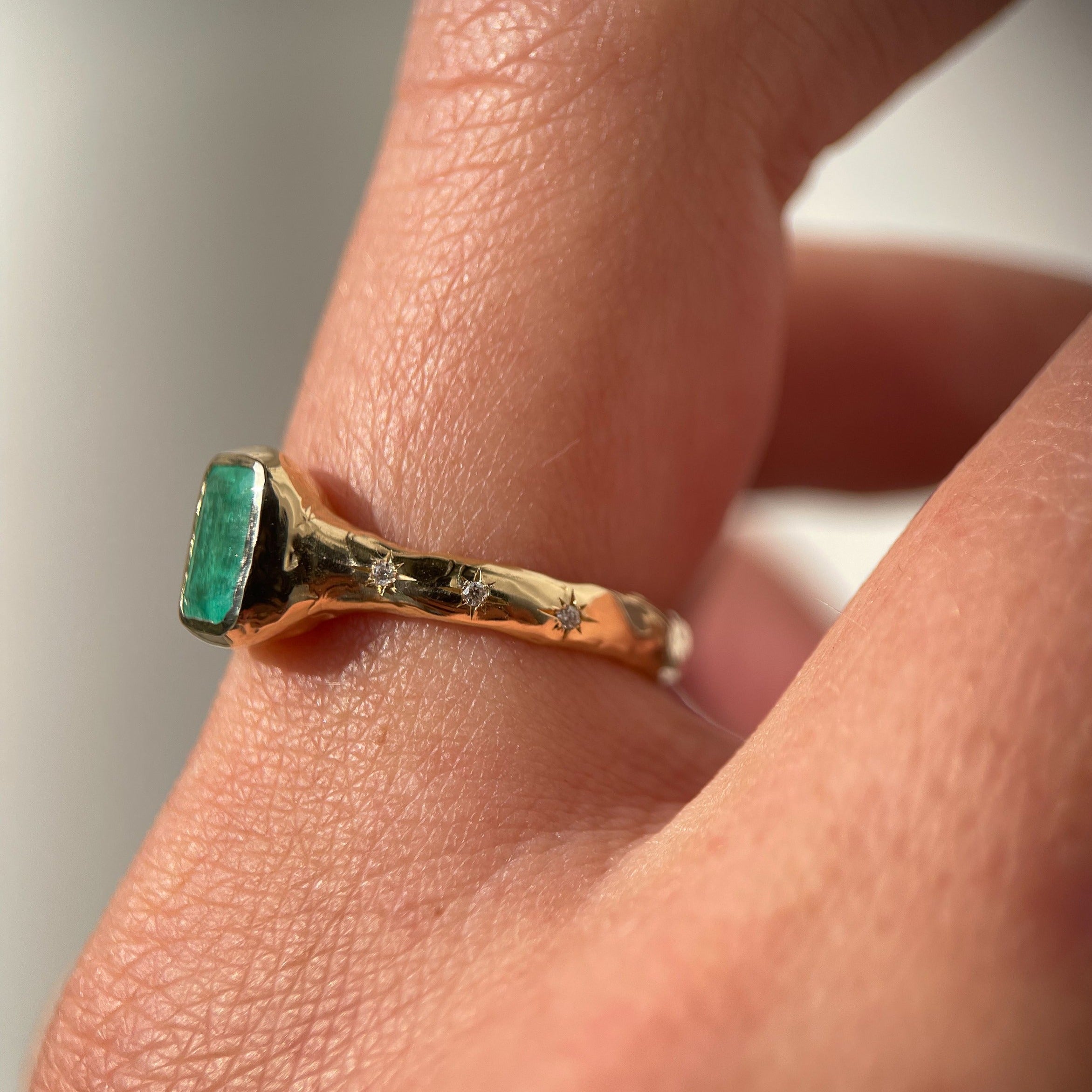 Side view of an emerald cut emerald ring is bezel set in 14k gold with a wide band that has multiple star set diamonds along the sides.