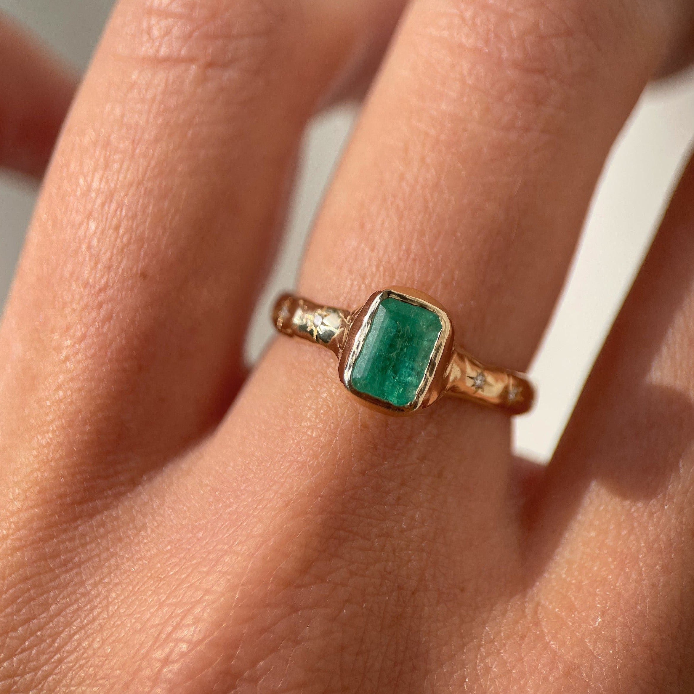 An emerald cut emerald ring is bezel set in 14k gold with a wide band that has multiple star set diamonds along the sides.