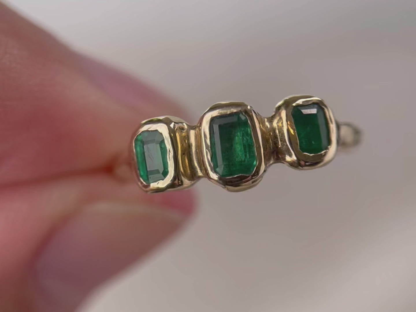 A close up video of a ring with three small emerald cut emeralds that are embedded into a 14k gold ring giving it an organic and handcrafted look.
