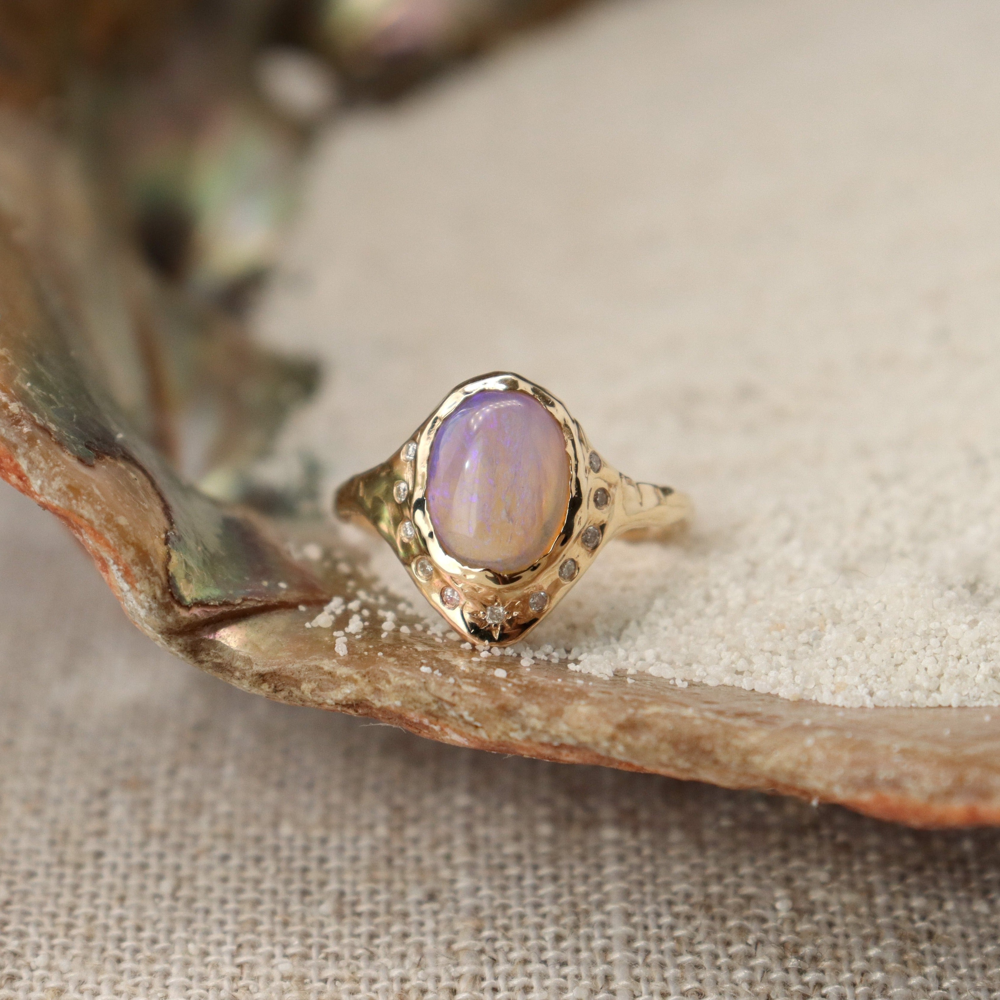 An oval opal is bezel set in 14k gold with diamonds set all around it, there is a star set diamond at the base, and the band is organically crafted..