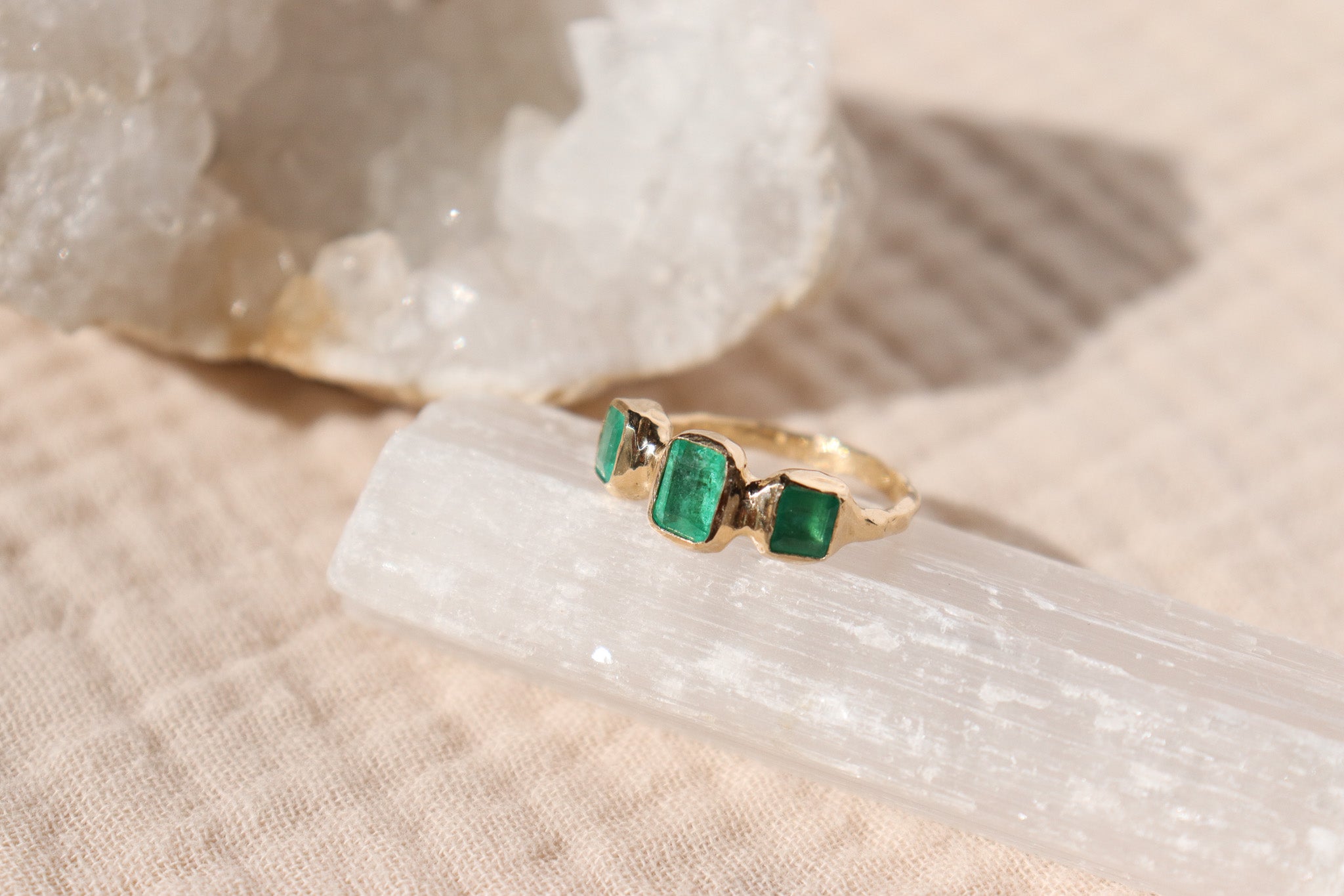 A 14k gold emerald trio ring is sitting atop a crystal on a cream surface.