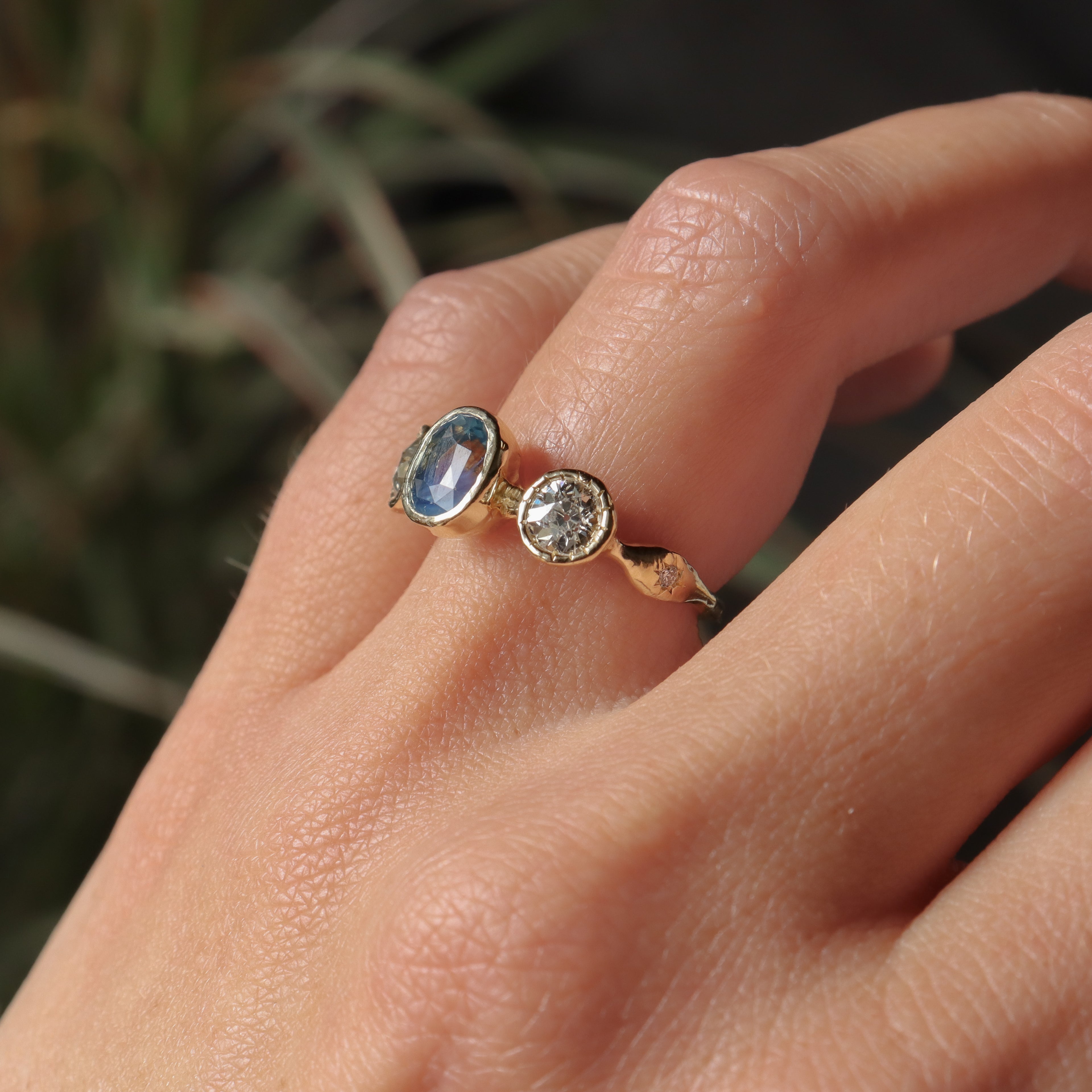 Close-up of a hand wearing a gold ring with gemstones against a blurred natural background