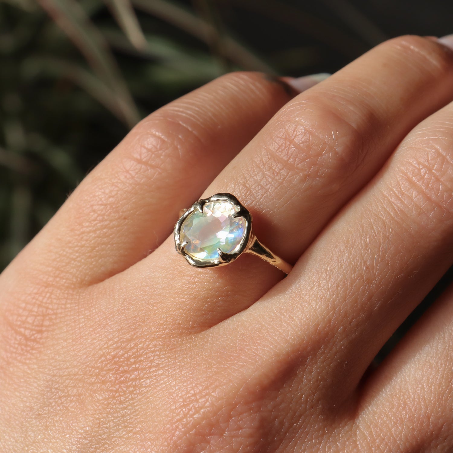 Close-up of a hand wearing a ring with a colorful gemstone against a blurred natural background