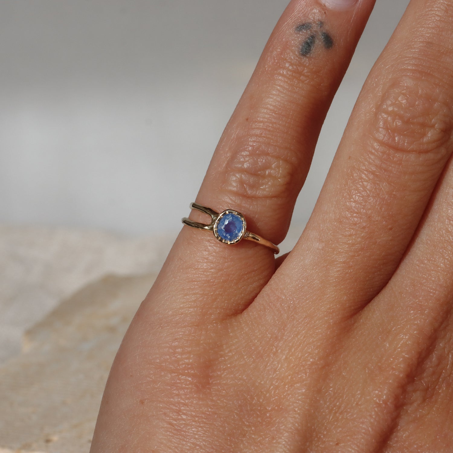 Hand wearing a gold ring with a blue gemstone on a neutral background