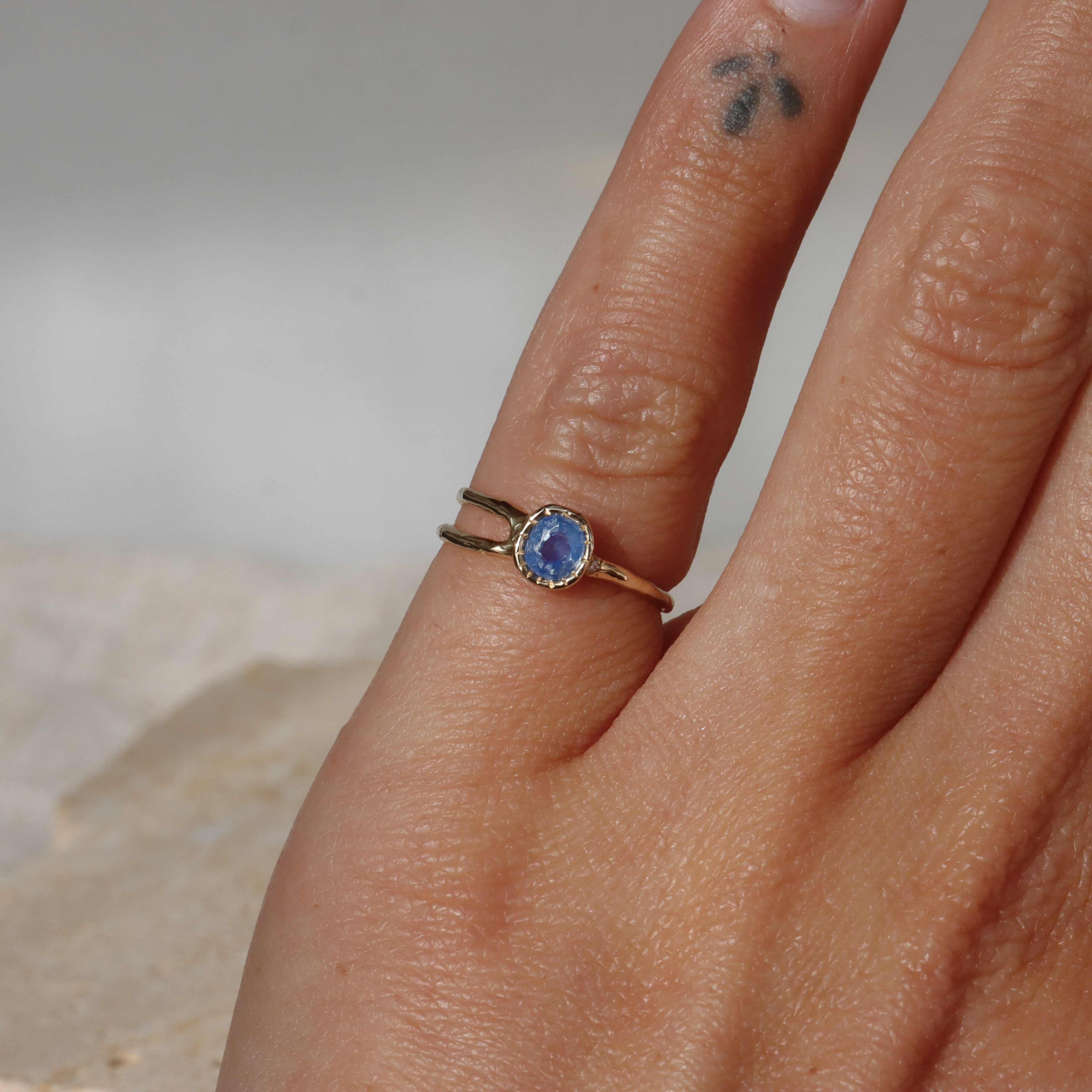 Hand wearing a gold ring with a blue gemstone on a neutral background