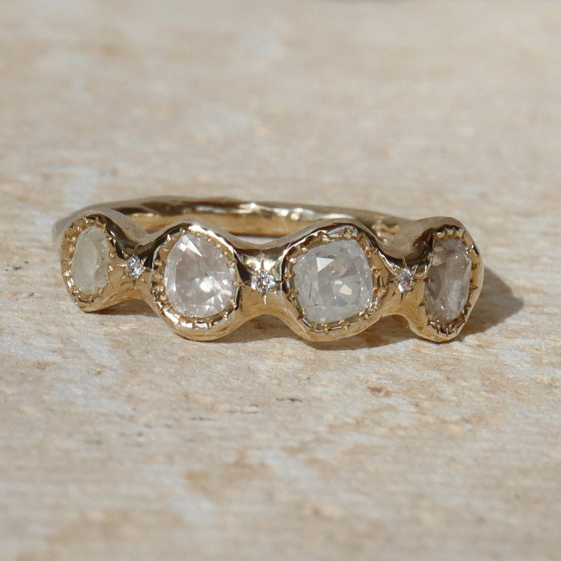 A gold band ring featuring five round-cut clear gemstones in a wavy row, photographed on a light beige stone surface.
