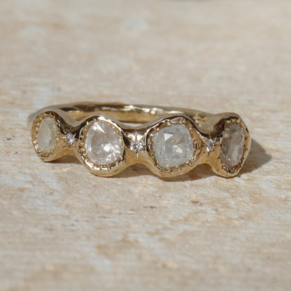 A gold band ring featuring five round-cut clear gemstones in a wavy row, photographed on a light beige stone surface.