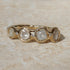 A gold band ring featuring five round-cut clear gemstones in a wavy row, photographed on a light beige stone surface.