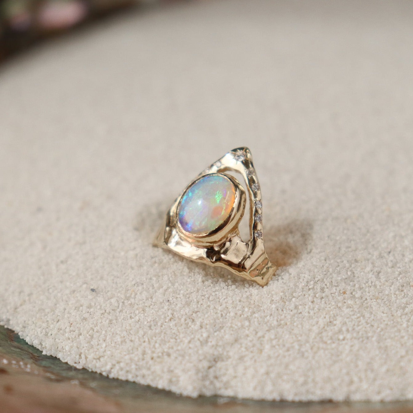 Side view of a ring featuring a bezel-set opal on a wide, organically shaped band, adorned with a crown-like V-shaped band encircling the opal and embellished with glistening diamond accents