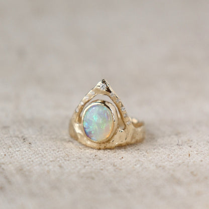 A ring featuring a bezel-set opal on a wide, organically shaped band, adorned with a crown-like V-shaped band encircling the opal and embellished with glistening diamond accents