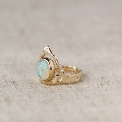 Side view of a ring featuring a bezel-set opal on a wide, organically shaped band, adorned with a crown-like V-shaped band encircling the opal and embellished with glistening diamond accents