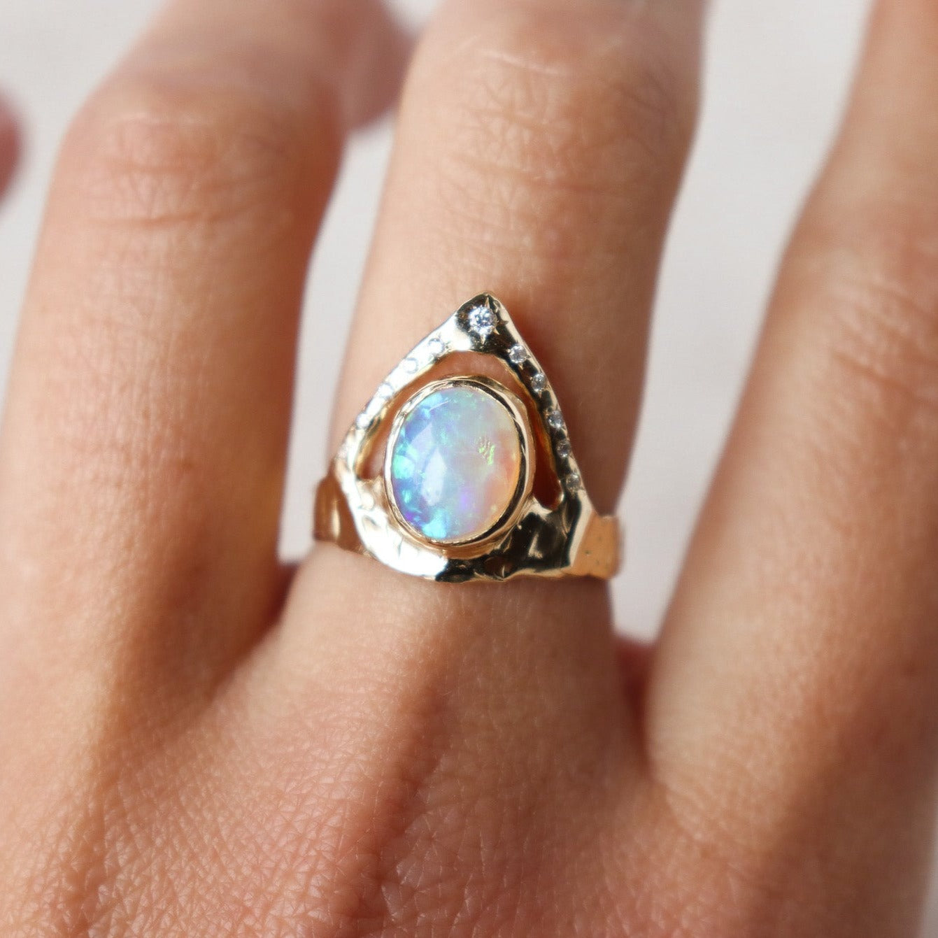 A ring featuring a bezel-set opal on a wide, organically shaped band, adorned with a crown-like V-shaped band encircling the opal and embellished with glistening diamond accents, shown on  a finger  for scale