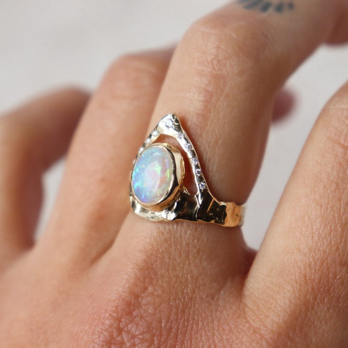 Side view of a ring featuring a bezel-set opal on a wide, organically shaped band, adorned with a crown-like V-shaped band encircling the opal and embellished with glistening diamond accents, shown on a finger for scale