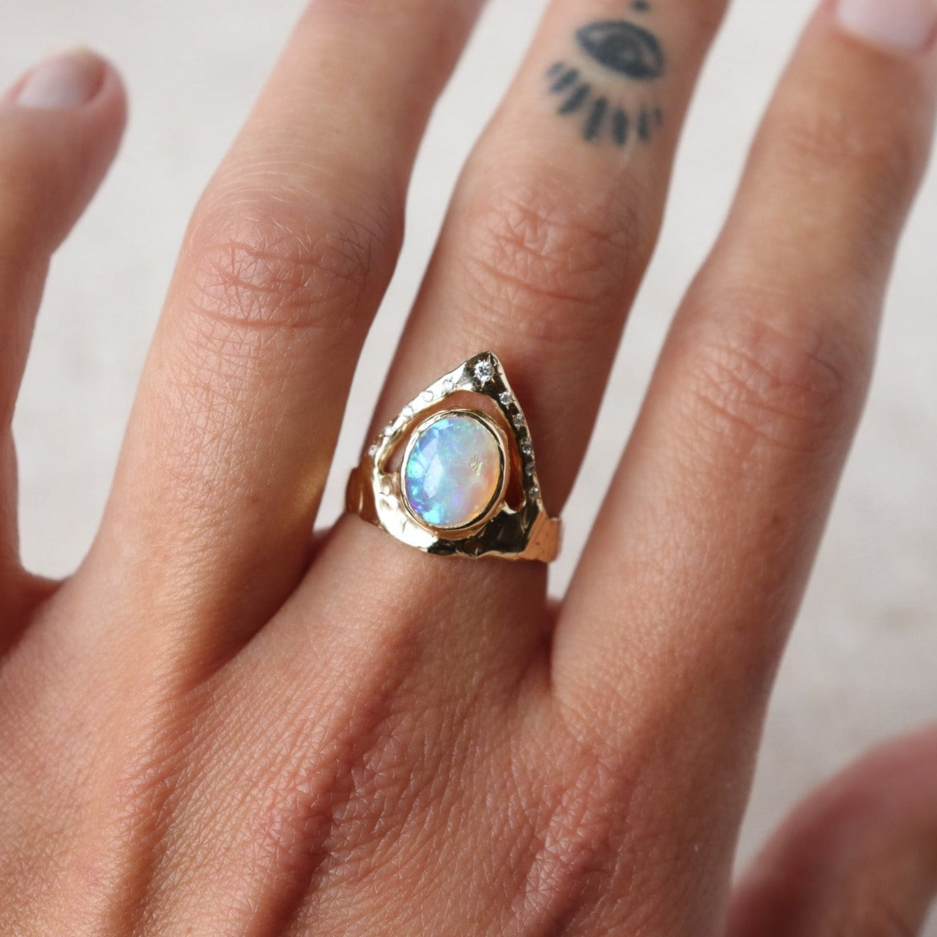 Stunning ring featuring a bezel-set opal on a wide, organically shaped band, adorned with a crown-like V-shaped band encircling the opal and embellished with glistening diamond accents, shown on  a finger for scale.