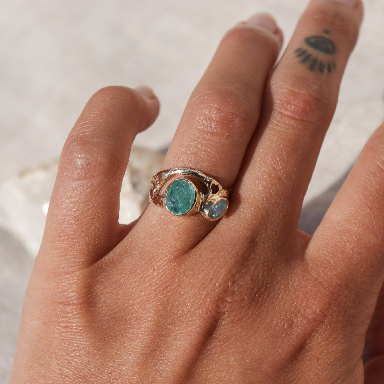 Organically shaped ring with a stunning Paraiba tourmaline and opal, bezel-set and accented with dazzling diamonds shown on a finger for scale.