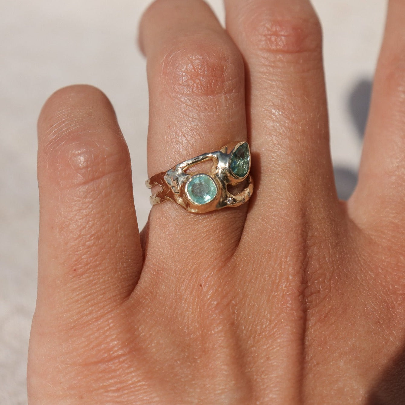 Exquisite ring featuring two vibrant Paraiba tourmalines elegantly bezel-set along a wide, organically shaped band worn on a finger and shown from the side to depict scale and show details.