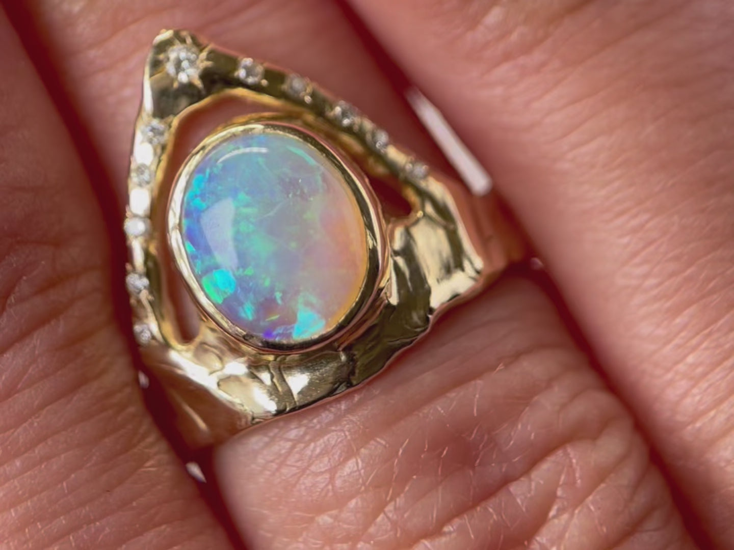 A close up video of a ring featuring a bezel-set opal on a wide, organically shaped band, adorned with a crown-like V-shaped band encircling the opal and embellished with glistening diamond accents