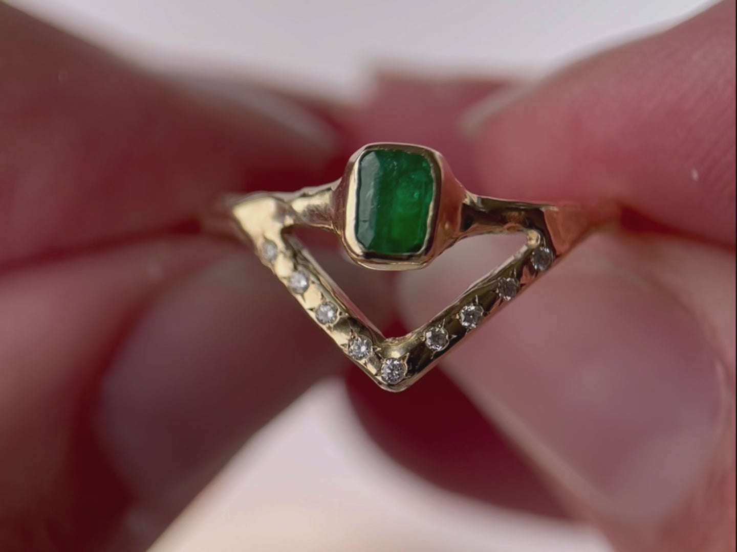 A close up video of an emerald ring with a bezel-set, rich green gemstone elegantly perched on a delicate, narrow band. Beneath it, a gracefully designed V-band features organically set diamonds, adding a touch of nature-inspired luxury to your jewelry collection.