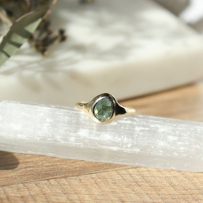 A rose cut blue green tourmaline is set in 14k  gold  into a signet style ring on a wooden table.
