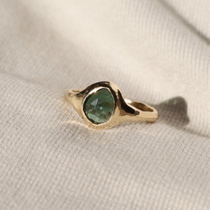 A rose cut blue green tourmaline is set in 14k  gold  into a signet style ring, on a white background.