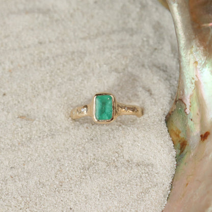 An emerald cut emerald ring is bezel set in 14k gold with a wide band that has multiple  star set diamonds along the sides.