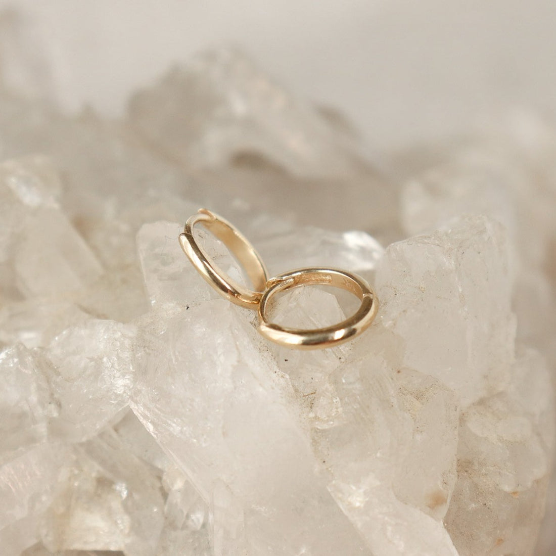 A pair of plain 14k gold huggie hoops are laid out on a crystal background.
