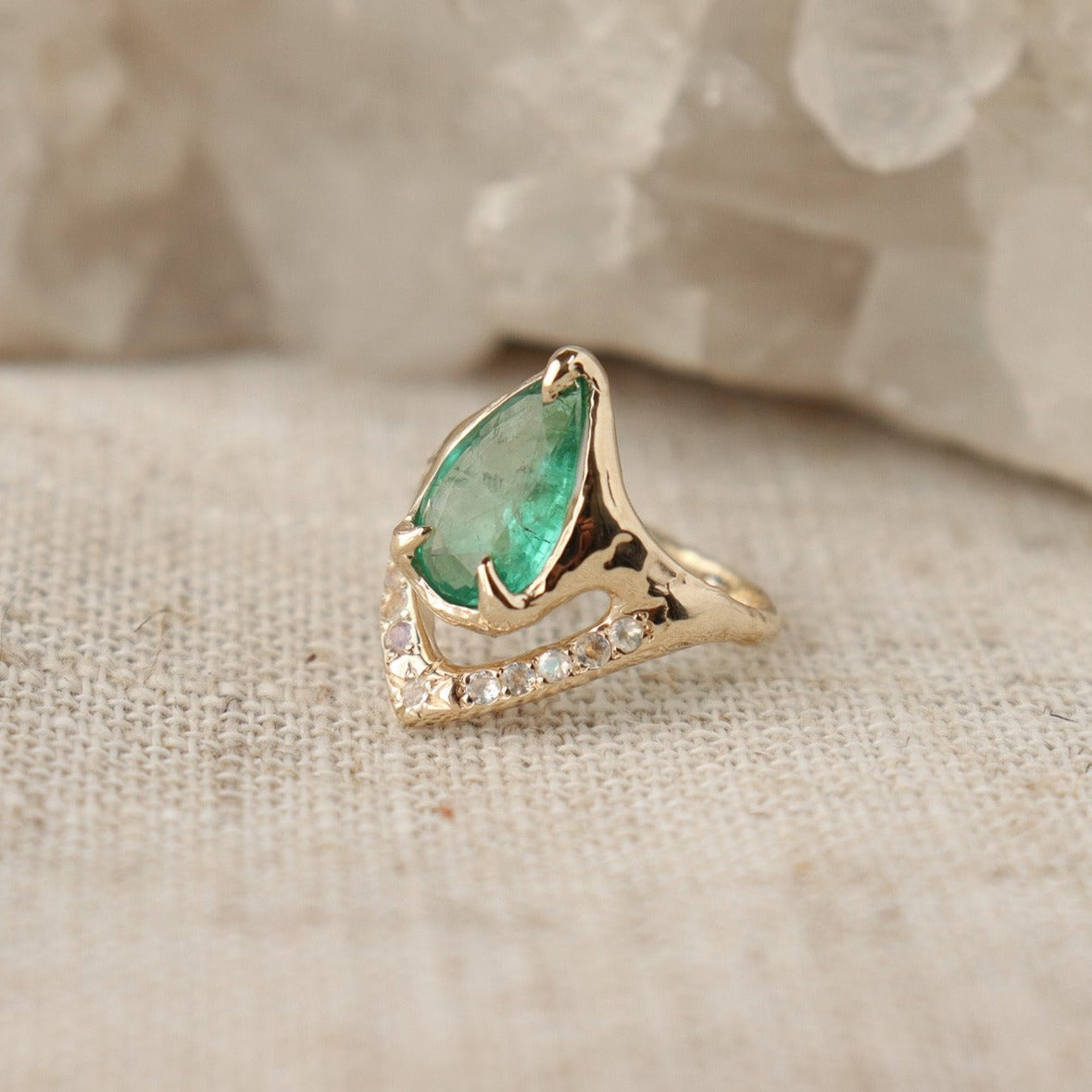 Close  up side view of a pear shaped emerald is set with prongs and has a V-shape band under the stone with moonstones.