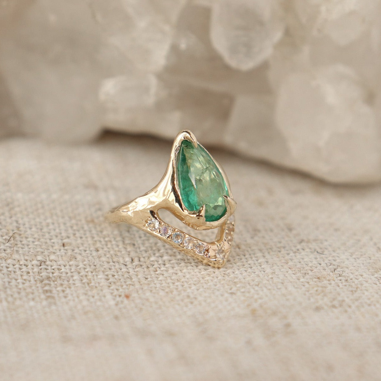 Side view of a pear shaped emerald is set with prongs and has a V-shape band under the stone with moonstones.