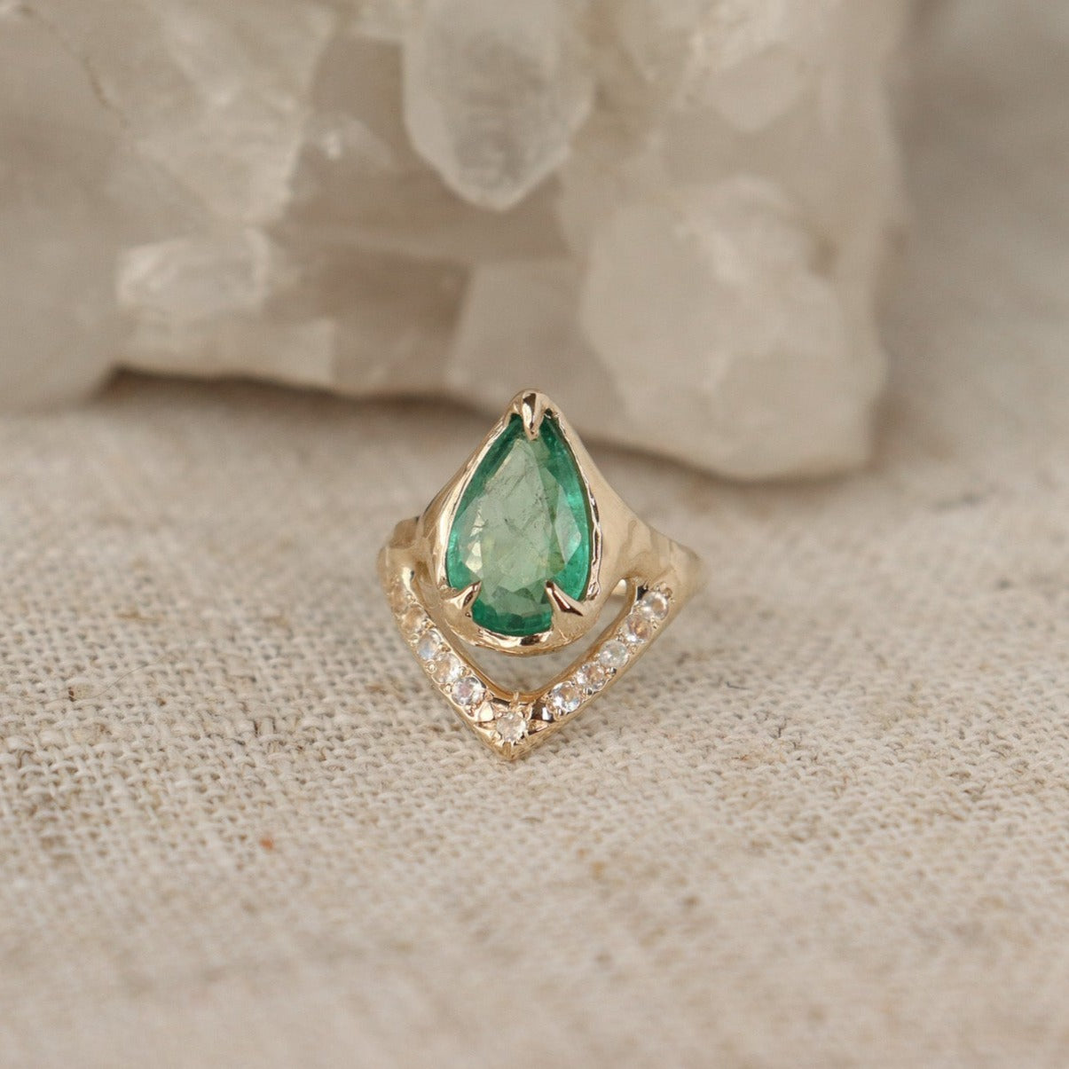 Front view of a pear shaped emerald is set with prongs and has a V-shape band under the stone with moonstones.