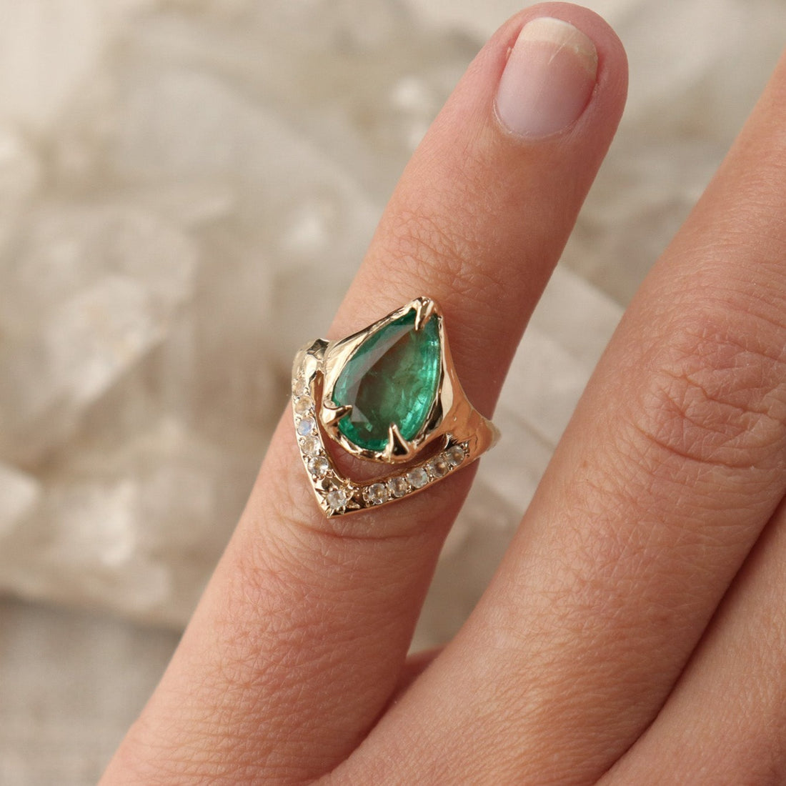 A pear shaped emerald is set with prongs and has a V-shape band under the stone with moonstones.
