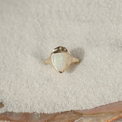 White triangle opal is bezel set in 14k gold with three star set diamonds, on a sand background.