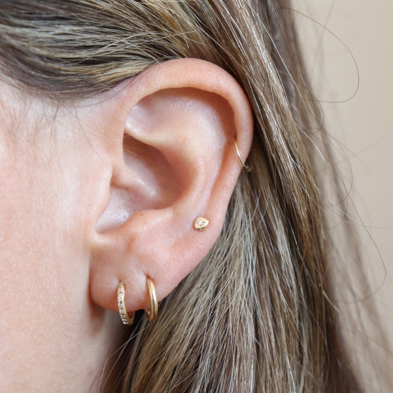 Multiple 14k gold earrings are worn, the top is a thin hoop, middle is a circle stud with a diamond set in the center, then two versions of a huggie hoop.