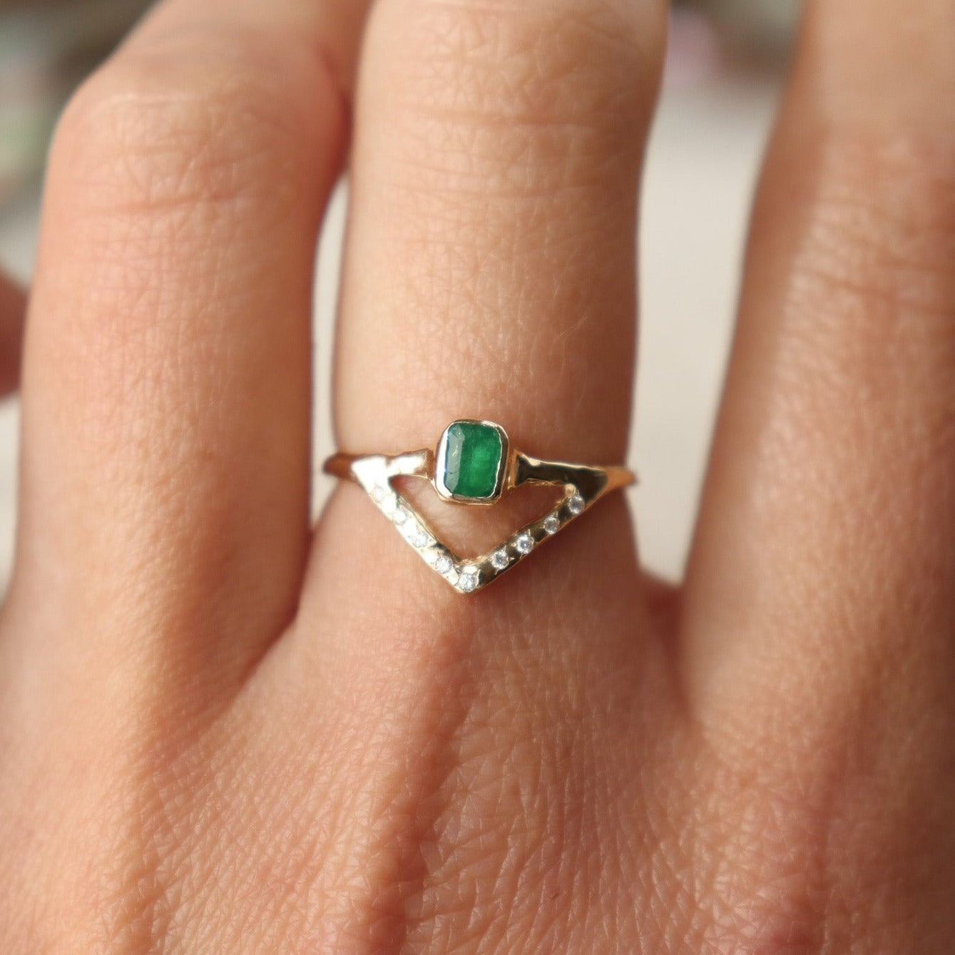 Stunning emerald ring with a bezel-set, rich green gemstone elegantly perched on a delicate, narrow band. Beneath it, a gracefully designed V-band features organically set diamonds, adding a touch of nature-inspired luxury to your jewelry collection. Worn on a hand to show scale.