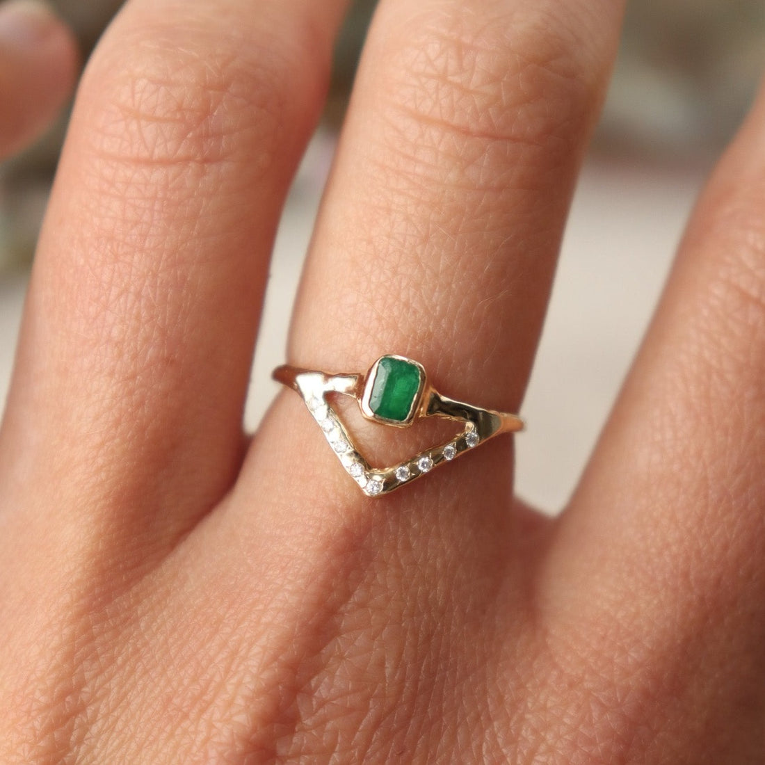 Stunning emerald ring with a bezel-set, rich green gemstone elegantly perched on a delicate, narrow band. Beneath it, a gracefully designed V-band features organically set diamonds, adding a touch of nature-inspired luxury to your jewelry collection. Worn on a hand to show scale.