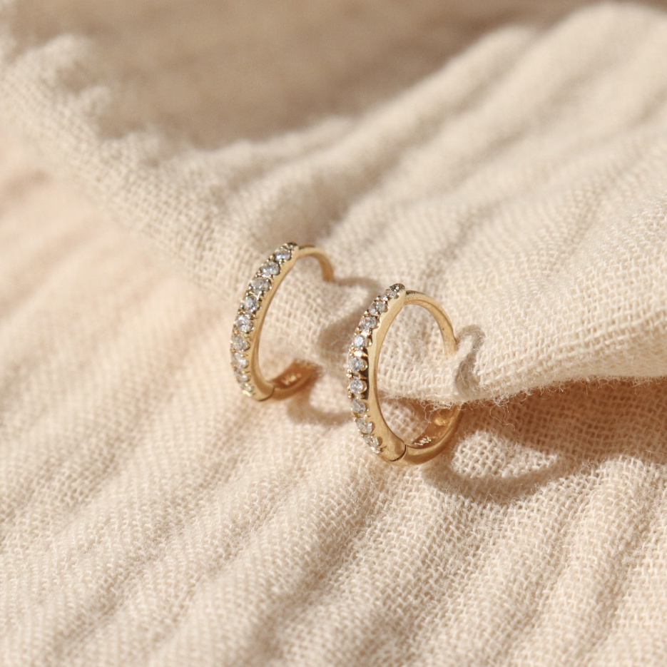 A 3/4 view of a pair of diamond pave huggie hoops positioned on cream fabric