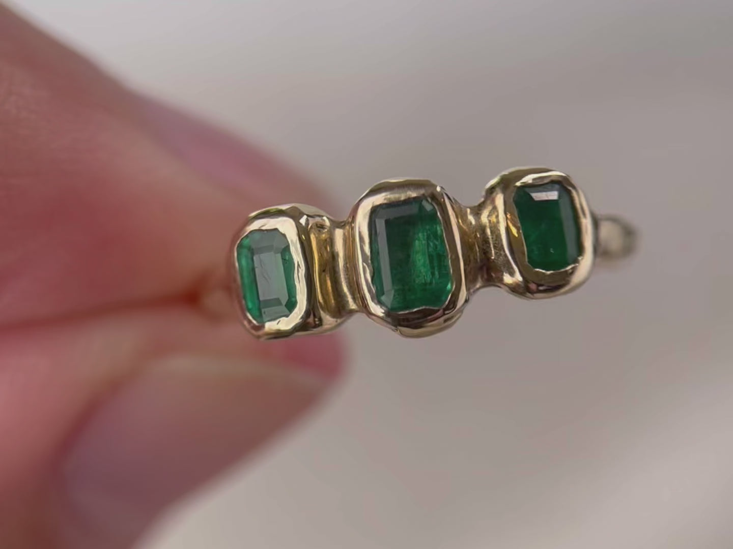 A close up video of  a ring with  three small emerald cut emeralds that are embedded into a 14k gold ring giving it an organic  and handcrafted look.
