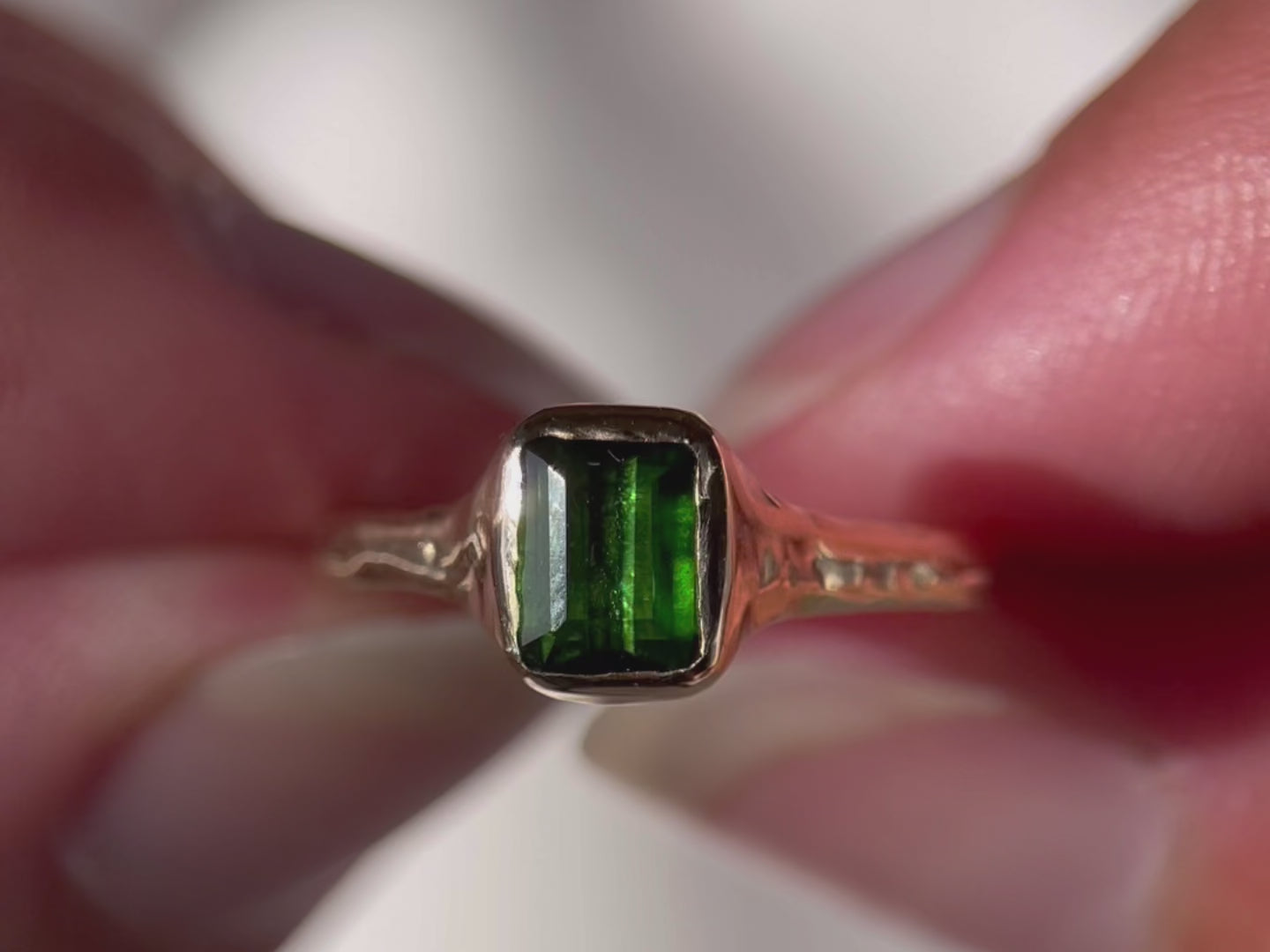 A close up video of an emerald cut dark green tourmaline is bezel set in 14k gold on an organic band.